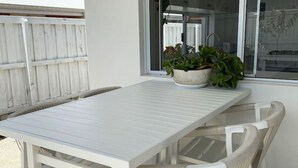 A white outdoor patio with a table and chairs, a window with a view of the backyard, and a potted plant on the windowsill.