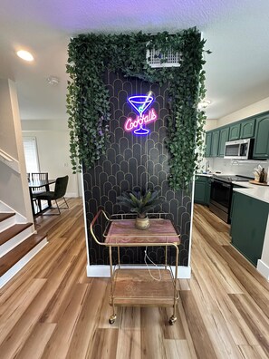 Una cocina moderna con armarios verdes, un letrero de neón con cócteles y suelo de madera.