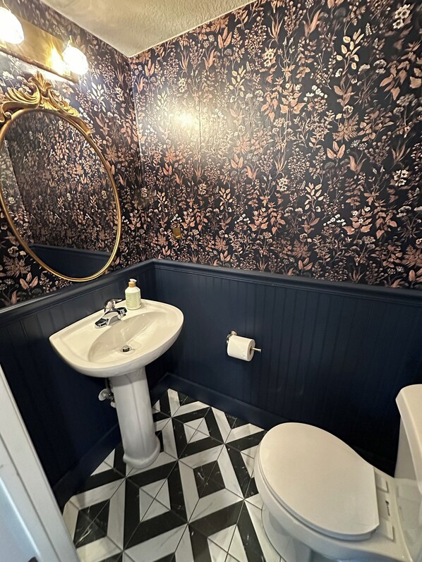 Un baño con lavamanos, inodoro y espejo decorativo. Las paredes están cubiertas de un papel tapiz floral, y el suelo presenta un patrón geométrico en blanco y negro.