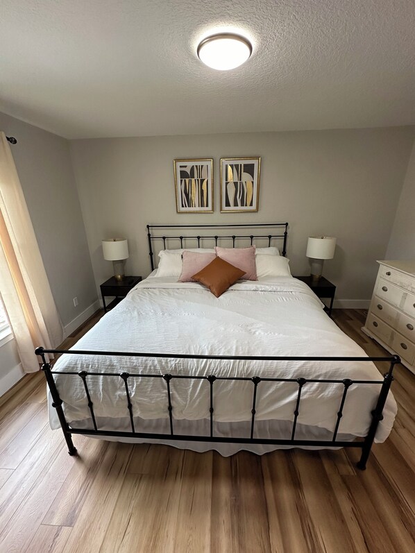 A bedroom with a metal bed frame, white bedding, two bedside tables with lamps, and two framed abstract artworks on the wall.