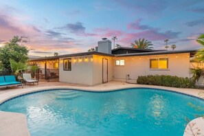 Una piscina con zona de patio, un edificio pequeño con luces colgantes y palmeras al fondo.