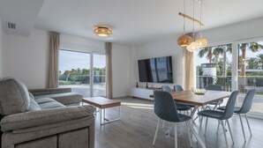 Un salon moderne comprenant un canapé, une table à manger, des chaises, un téléviseur et de grandes fenêtres donnant sur des palmiers.