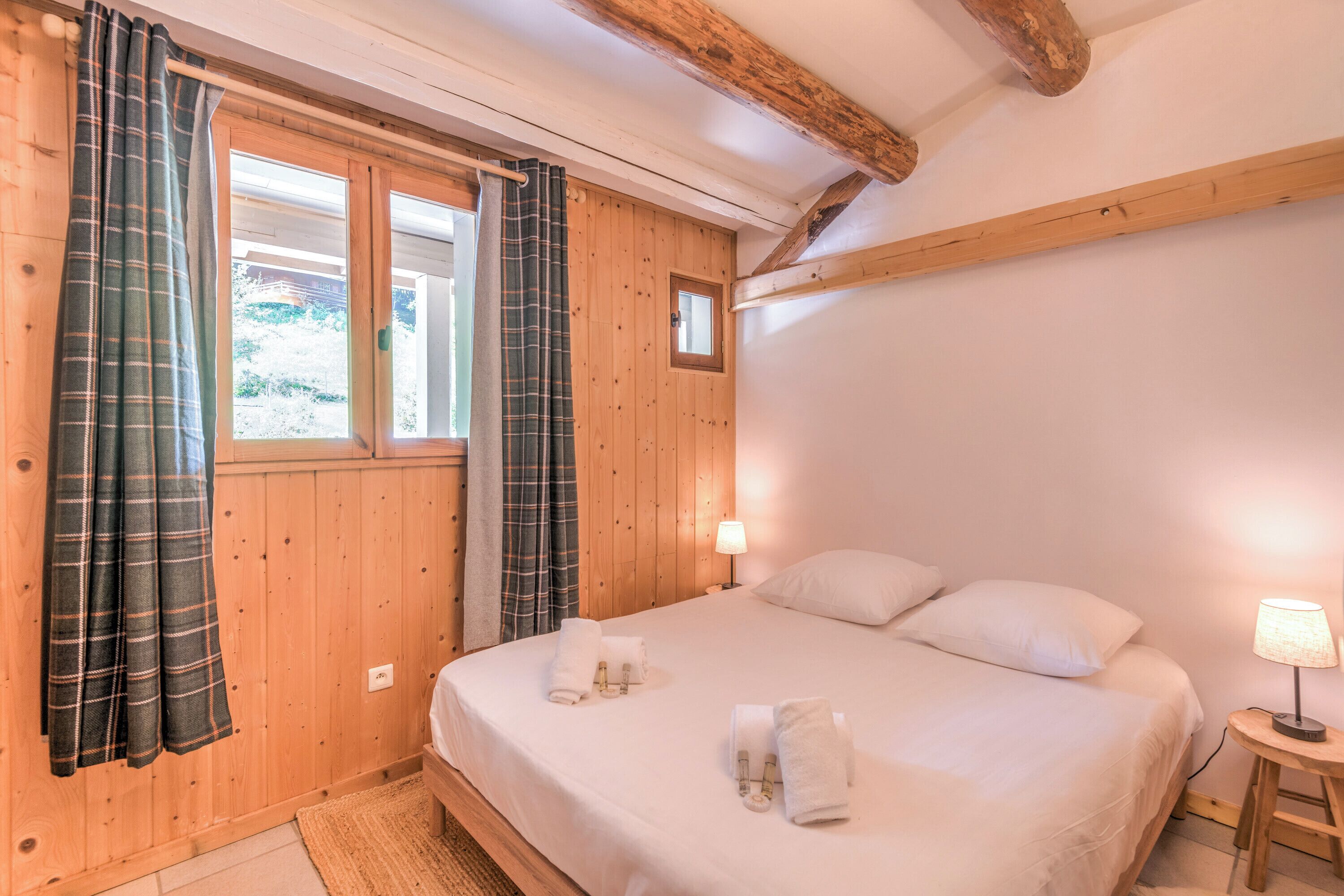 A wooden-paneled bedroom with a bed, two pillows, a nightstand with a lamp, and a window with curtains.