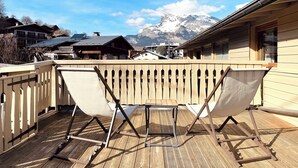 A wooden deck with two white folding chairs, a wooden railing, and a view of snow-capped mountains in the background.