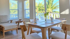 A spacious dining area with a high ceiling, wooden furniture, and large windows offering a view of palm trees. - NEW 2 bdrm Sunrise Villa on 1+ acre tropical garden directly on Hoopers Beach (George Town)