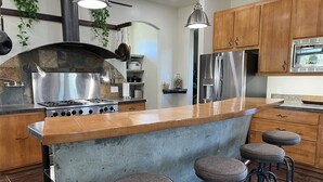 Una cocina con una isla central, taburetes de bar, electrodomésticos de acero inoxidable, armarios de madera y plantas colgantes.
