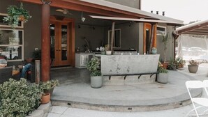 Un patio techado al aire libre con zona de estar de concreto, plantas en macetas y una puerta de madera que da a la casa.