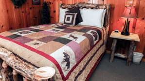 A wooden-paneled room with a bed featuring a quilt with animal prints, a nightstand with a lamp, and a wall-mounted decoration.