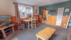 A cozy room with a wooden dining table, chairs, a television on a wooden stand, and a large window with curtains.