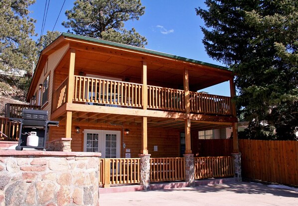 A two-story wooden cabin with a balcony, stone foundation, and a wooden fence. - Walk to Downtown from Spacious Home with Hot Tub #3383 (Estes Park)