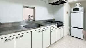 A modern kitchen with white cabinets, a stainless steel countertop, a sink, a microwave, and a refrigerator. - Dai3kuboi 304 (Hiroshima)