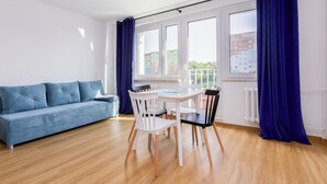 Una sala de estar moderna con un sofá azul, una mesa de comedor con sillas, ventanas amplias con cortinas azules y vistas a edificios.