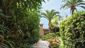 A stone pathway leading to a stone building with a tiled roof, surrounded by lush greenery and flowering plants.