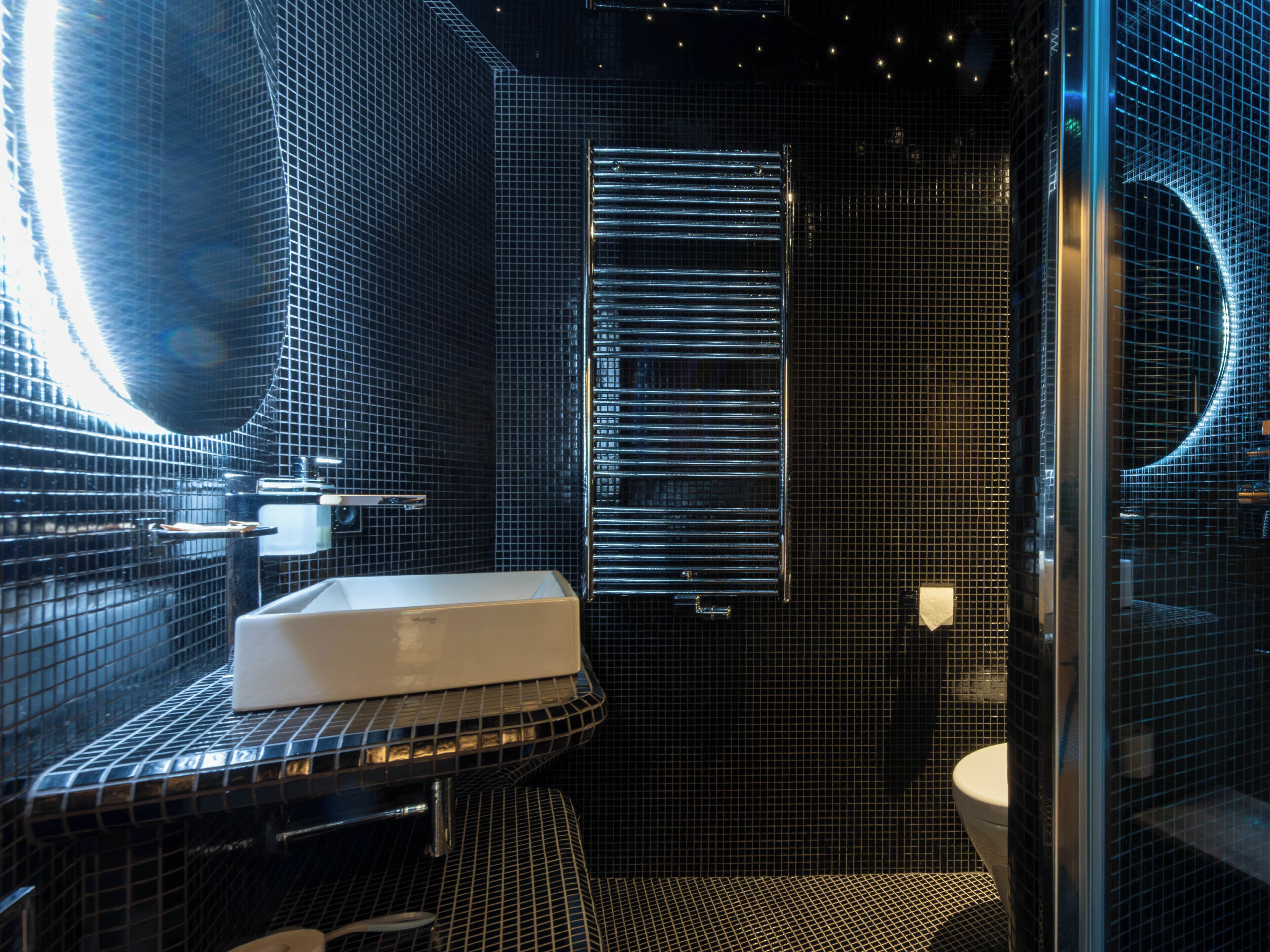 Une salle de bain moderne avec un lavabo blanc, une douche en verre et un mur recouvert de carreaux noirs.
