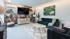 A modern living room with a flat-screen TV, a green sofa, a white coffee table, and a tropical-themed wall mural.