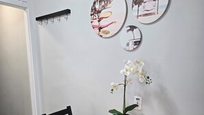 Zona pranzo con tavolo rotondo in legno, sedie nere e un'orchidea in vaso. Sono presenti ganci a parete e un'opera d'arte circolare raffigurante scene di spiaggia.