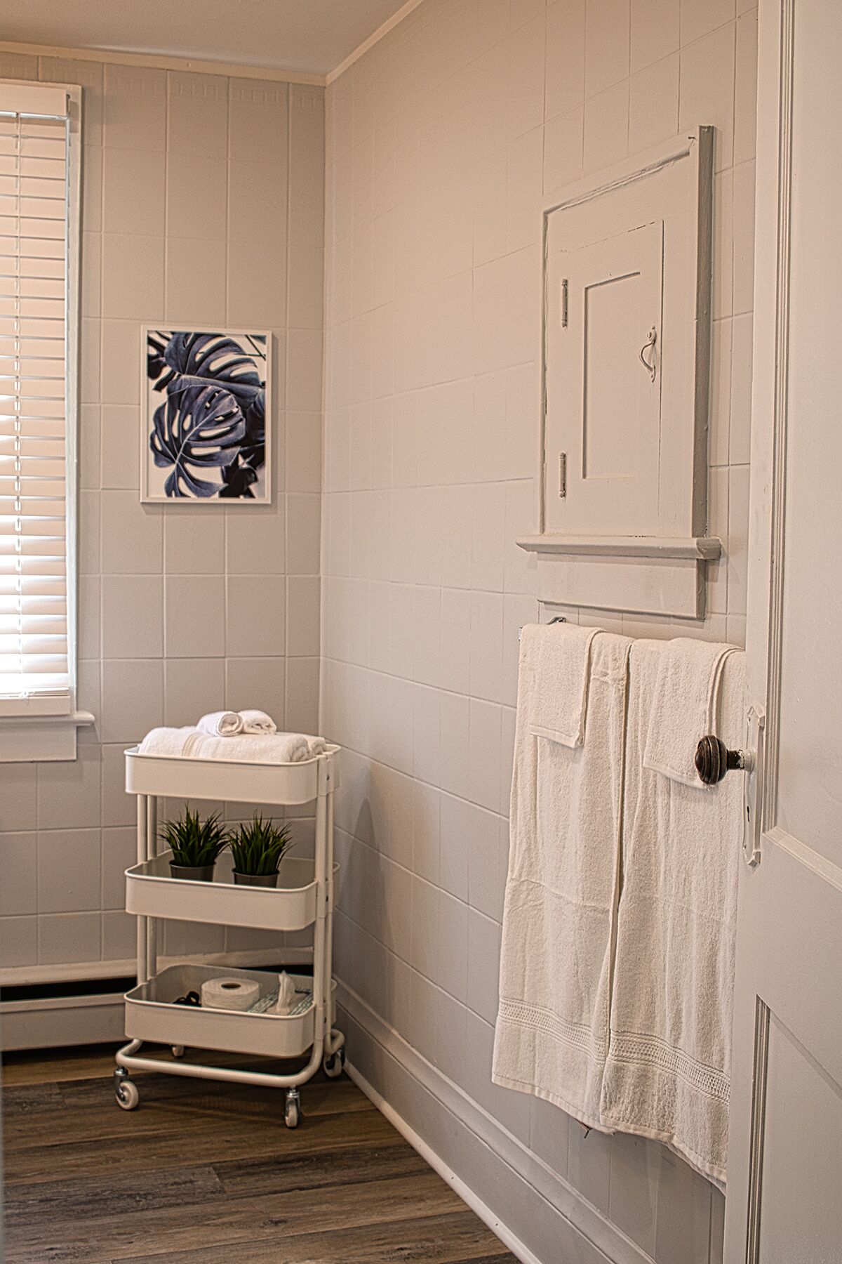 A bathroom with a white tiled wall, a door with a towel hanging on it, a white cart with towels and plants, and a framed picture on the wall.