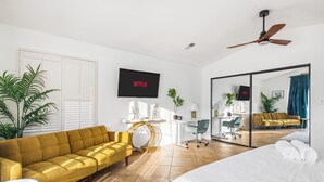 Quarto de hotel moderno com sofá amarelo, uma cama grande, uma TV exibindo 'NETFLIX', um ventilador de teto e um espelho grande refletindo o quarto.