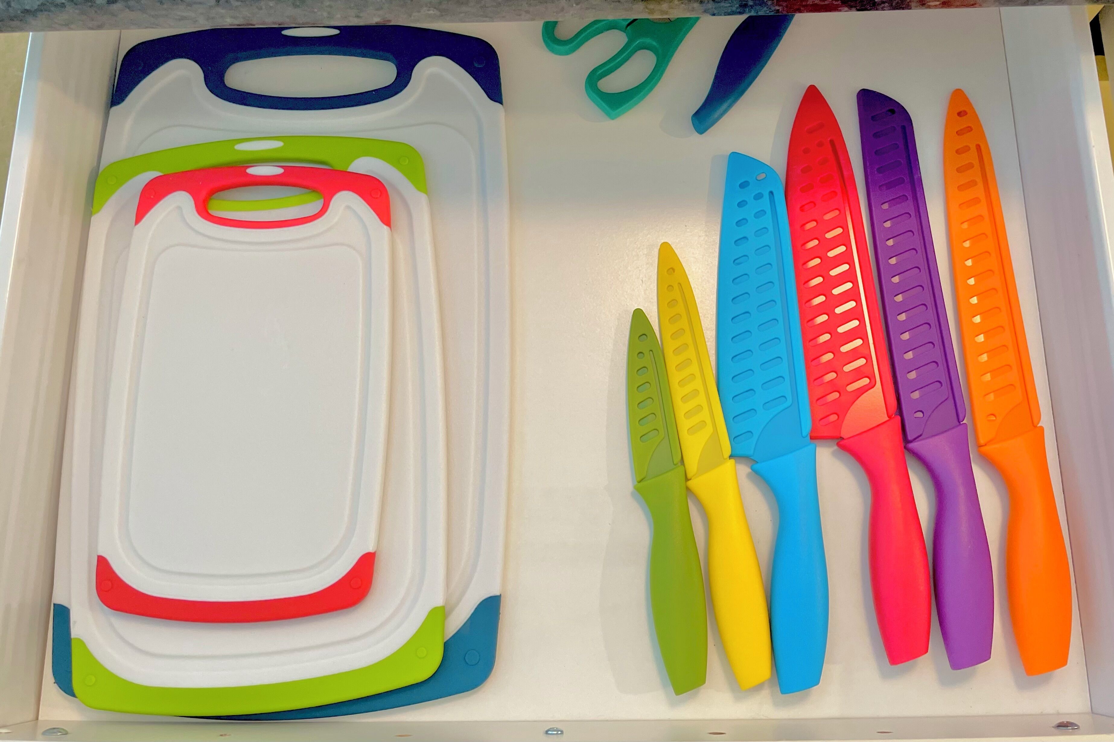 Un cajón con varios utensilios de cocina, incluyendo una tabla de cortar de plástico blanco y cuchillos de plástico de colores.