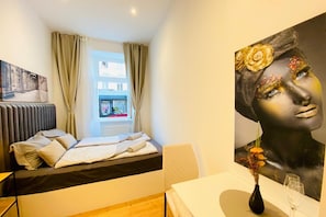 Una habitación de hotel con una cama, una ventana con cortinas, un cuadro en la pared y un florero con flores sobre una mesa.