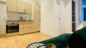 A compact kitchen with modern appliances, wooden cabinets, and a dark green sofa in a bright room with white walls. - Ottakring Gem/Cozy Room /w Breathtaking City Views (Vienna)