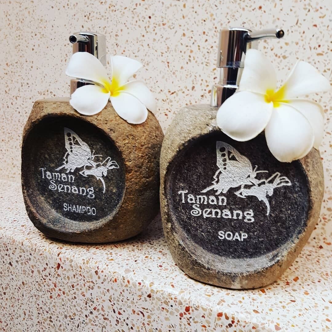 Two stone containers with engraved designs and white flowers on top, labeled 'Taman Senang Shampoo' and 'Taman Senang Soap'.