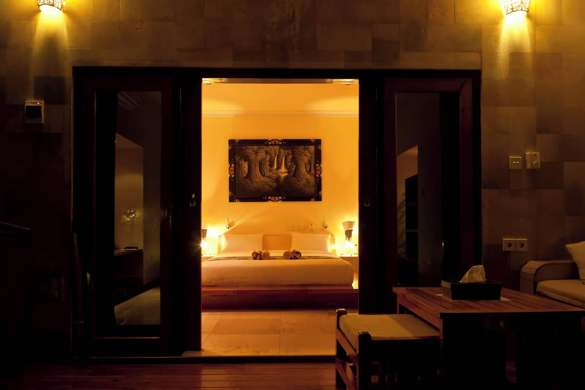 A hotel room with a bed, bedside tables, a painting on the wall, and a view into the room.