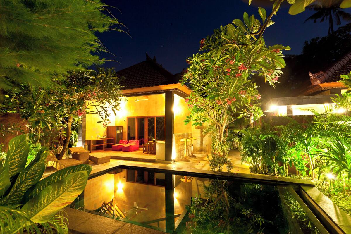 A modern house with a glass pool, surrounded by lush greenery and illuminated by warm lighting.