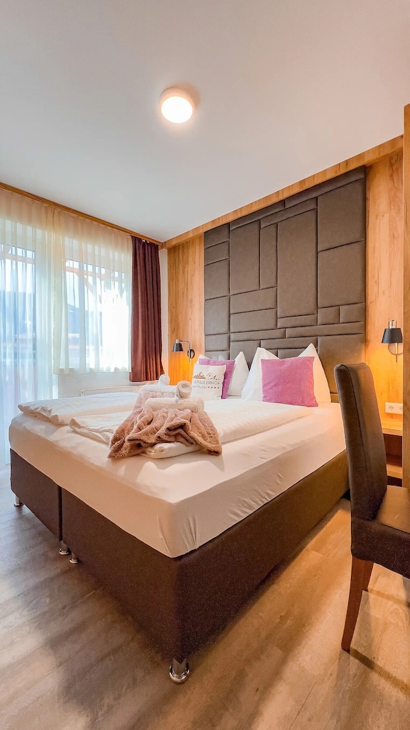 A hotel room with a large bed, a wooden headboard, a desk with a chair, and a window with curtains.