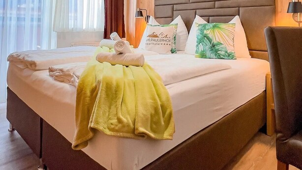 Ein Hotelzimmer mit einem großen Bett, einem Kopfteil mit geometrischem Muster, einer Wandpaneele aus Holz und einem Fenster mit transparenten Vorhängen.