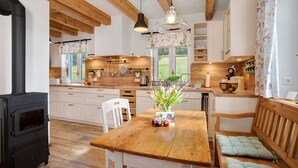 Une cuisine avec des poutres en bois, un poêle noir, des armoires blanches et une table en bois sur laquelle est posé un vase de fleurs.
