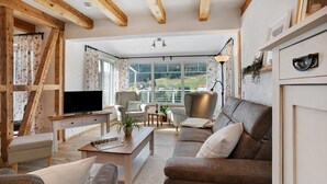 Un salon comprenant un téléviseur, un canapé, des fauteuils, une table basse et offrant une vue sur un paysage grâce à de grandes fenêtres.