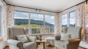 Une pièce avec deux fauteuils à motifs, une petite table sur laquelle se trouvent une bouteille et un verre, et de grandes fenêtres donnant sur un paysage verdoyant.