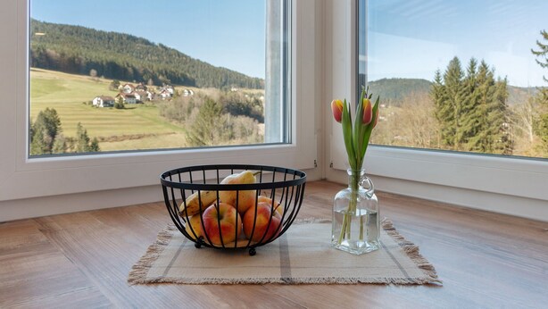 A window view of a rural landscape, a black wire fruit basket with apples and pears, and a clear vase with tulips on a wooden floor.