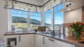 A kitchen with white cabinets, a wooden countertop, a coffee maker, a compost bin, and a view of a green landscape through large windows.