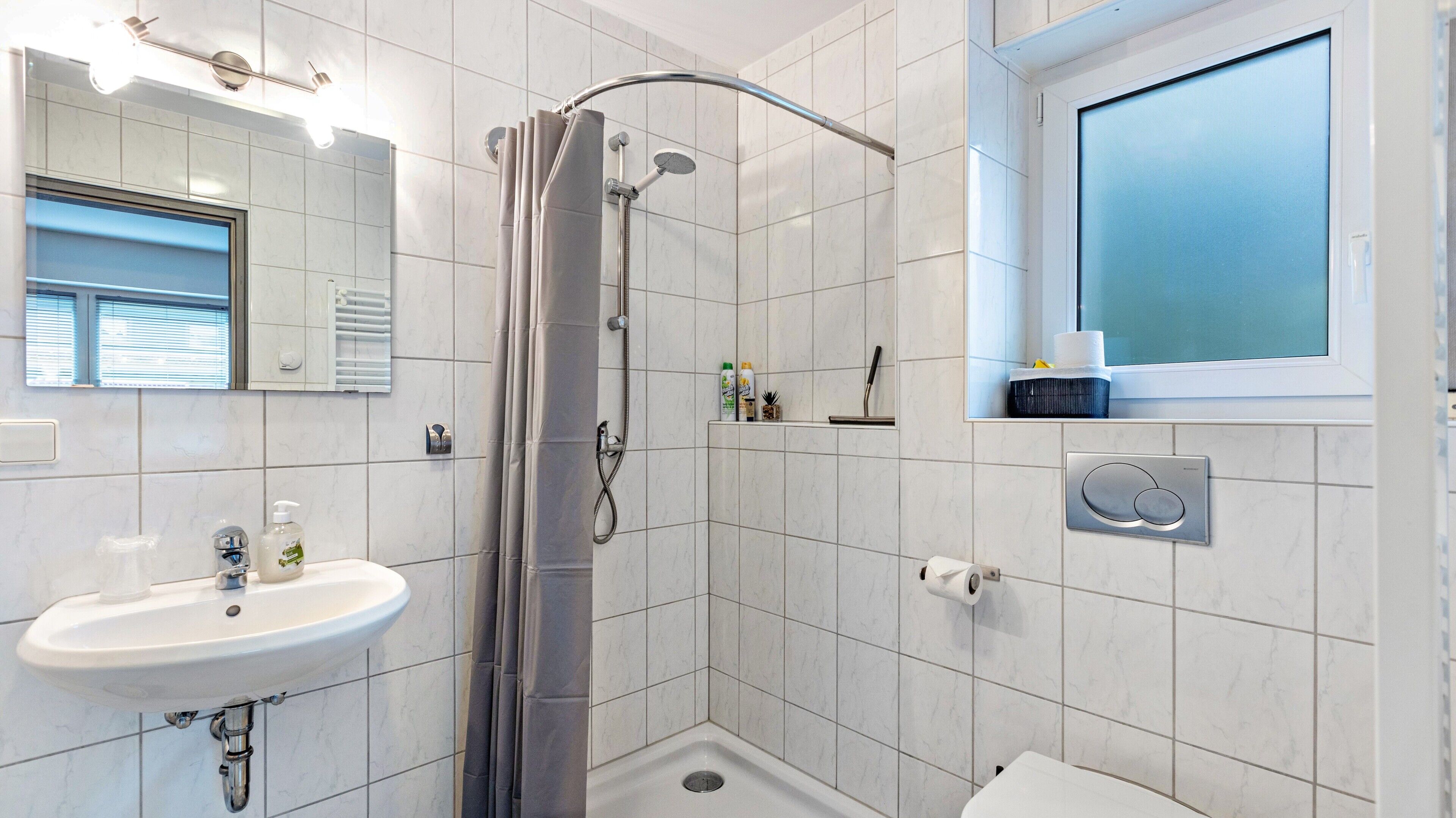 A modern bathroom with a shower, sink, toilet, and a window with blinds.