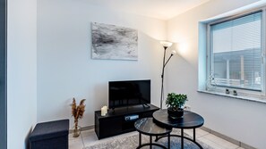 Un salon moderne avec un téléviseur à écran plat, une lampe sur pied, une petite table, un vase contenant des plantes séchées et une fenêtre avec des stores.