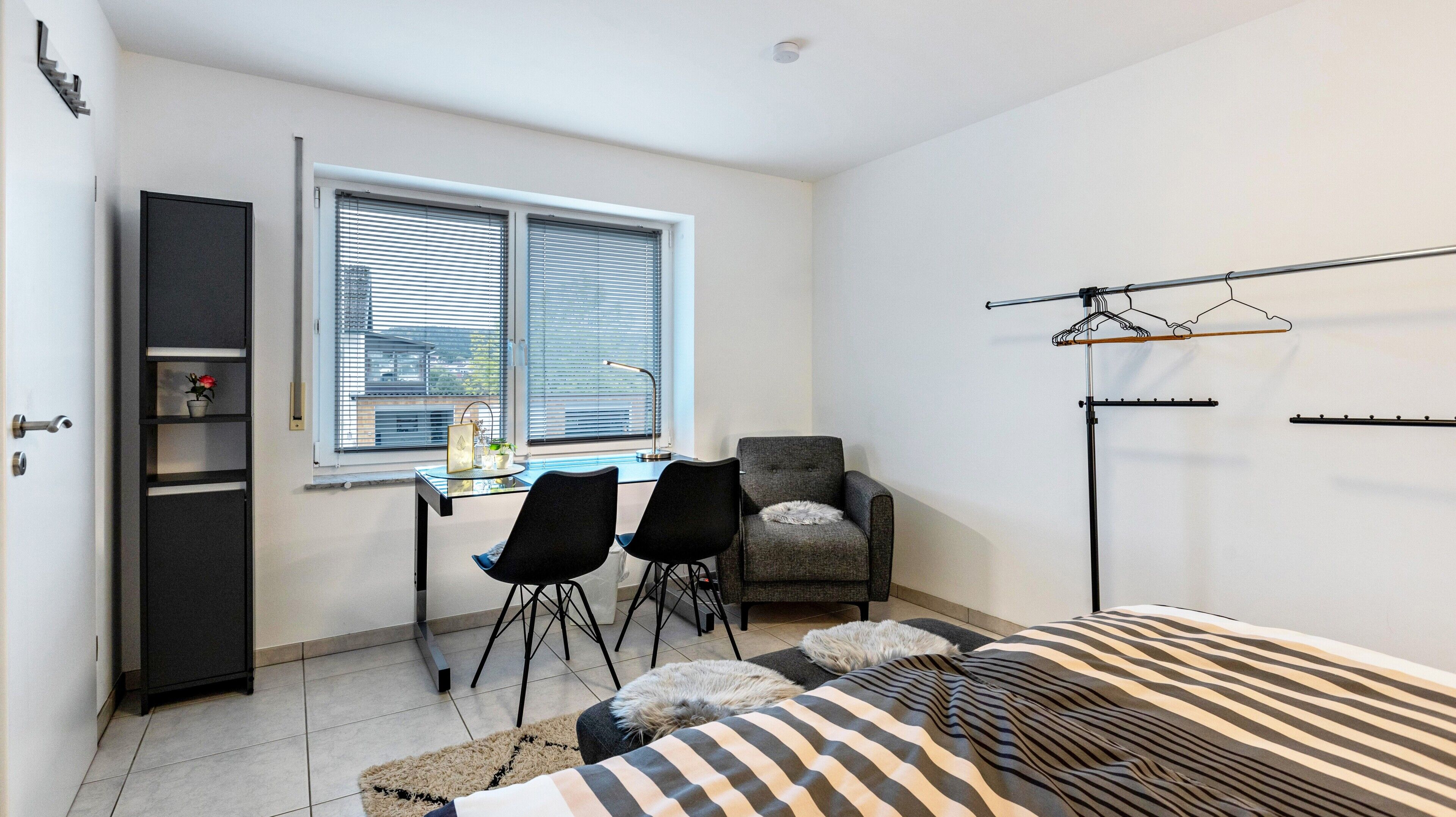 A small, modern room with a desk, two chairs, a grey armchair, a bed with striped bedding, a coat rack, and a window with blinds.