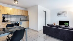 A modern kitchen with wooden cabinets, a dining table with chairs, a sofa, a TV, and a door.
