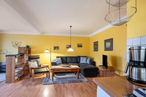 Un salon comprenant un canapé, un fauteuil, une table basse, une bibliothèque, un luminaire suspendu et un coin cuisine avec une machine à café.
