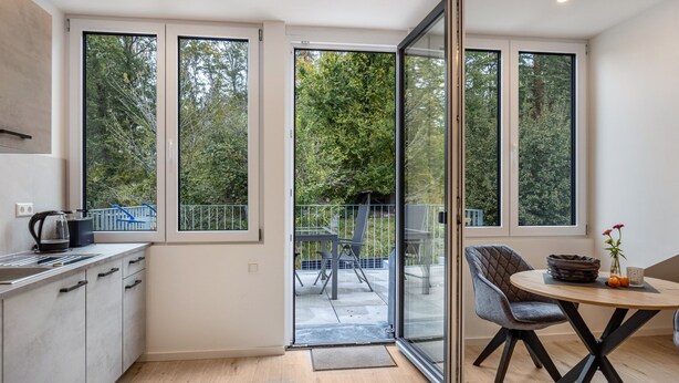 Eine moderne Küche mit Kaffeemaschine, ein Essbereich mit Tisch und Stühlen sowie große Fenster mit Blick auf Bäume.