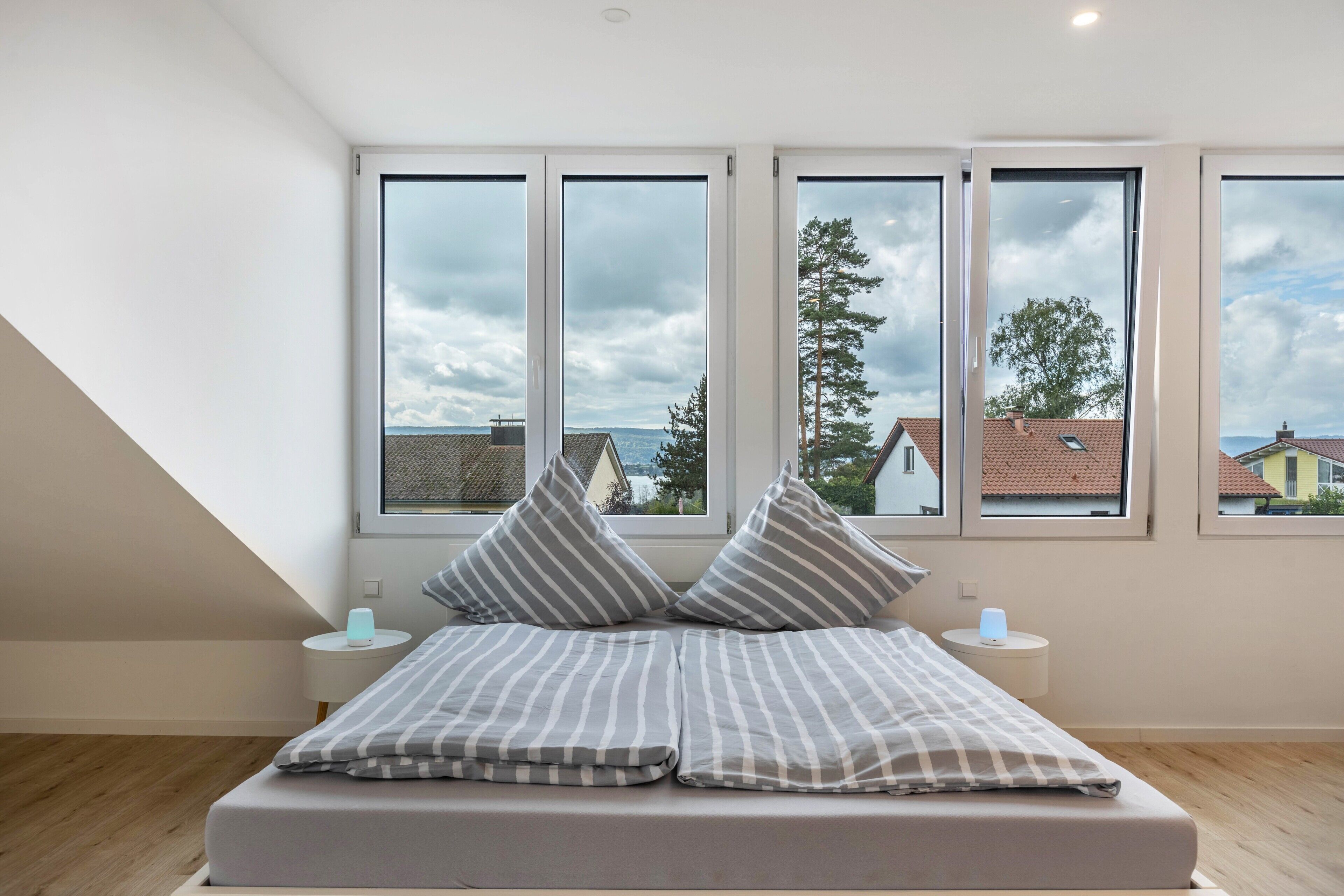 A bedroom with a bed, pillows, bedside tables, and large windows with a view of houses and trees.