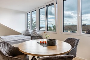 A modern room with a round table, cushioned chairs, a bed with striped pillows, and large windows offering a view of the sky and buildings.
