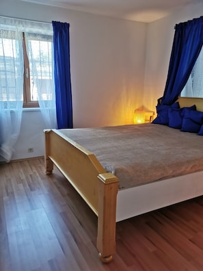 A bedroom with a wooden bed, blue curtains, a window with sheer curtains, and a bedside lamp.