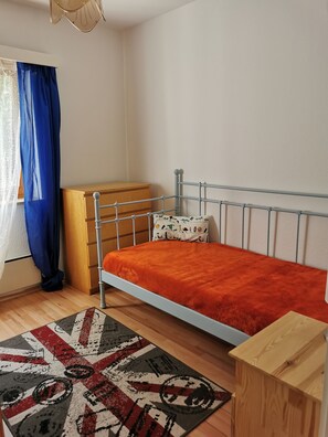 Un dormitorio con estructura de cama de metal, cómoda de madera, alfombra estampada y una ventana con cortinas azules.