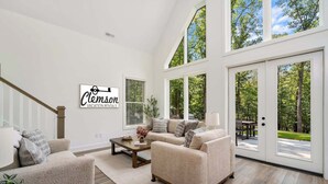 A spacious living room with a large window, a staircase, and a sign for Clemson Vacation Rentals. - Clemson Cove - Beautiful new construction on outskirts of Clemson! (Central)