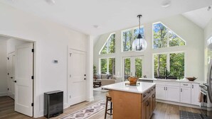A spacious kitchen with large windows, a central island, and a cozy seating area visible through the door. - Clemson Cove - Beautiful new construction on outskirts of Clemson! (Central)