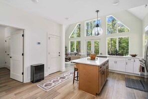 A spacious kitchen with large windows, a central island, and a cozy seating area visible through the door.