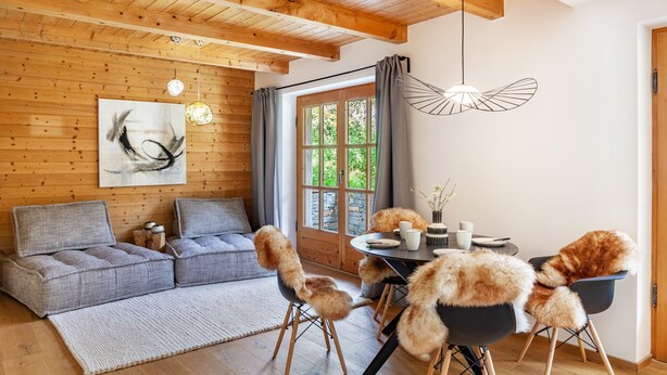 Una habitación acogedora con techo de madera, un sofá gris, una mesa de comedor con sillas y un ventanal con cortinas.