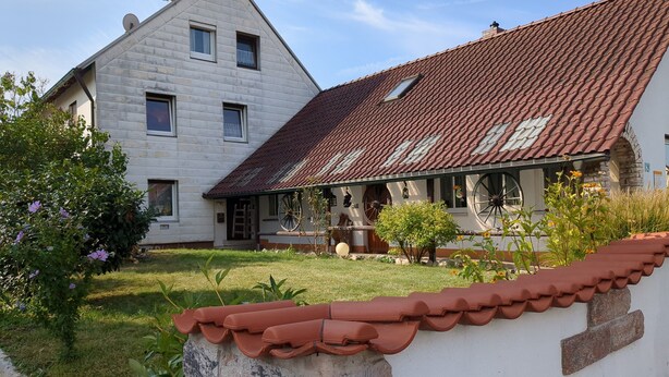 A two-story house with a red-tiled roof, a small porch, and a garden with flowers and shrubs.